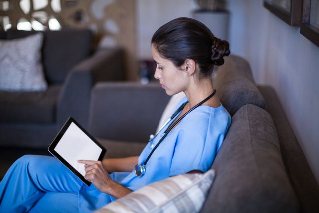 Nurse in blue scrubs sitting on a couch in a hospital lounge, using a digital tablet. Ideal for illustrating modern healthcare, technology in medicine, and the role of nurses in patient care. Suitable for healthcare websites, medical blogs, and educational materials about nursing and healthcare technology.