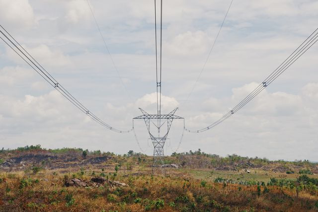 This depicts high tension power lines extending across an arid landscape. Clouds scatter across the sky in the background while the utility tower supports the intricate web of electrical cables. Ideal for uses in contexts related to energy, infrastructure development, rural electrification or environmental impact of utility projects.