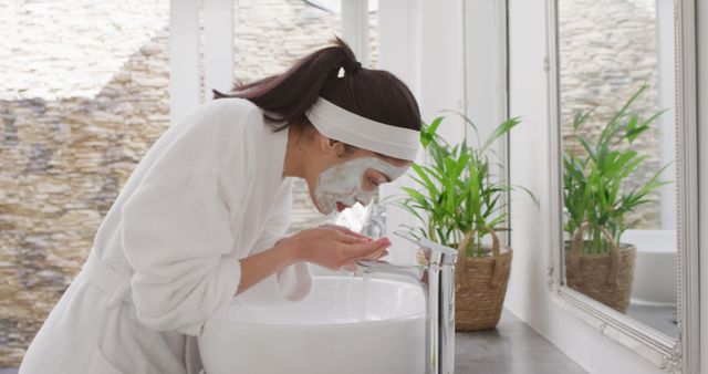 Image shows a woman rinsing off a face mask in a modern bathroom while wearing a white bathrobe and headband. Large mirror and plants in baskets can be seen in the background. Ideal for skincare products, self-care routines, health and wellness blogs, or bathroom decor inspiration.