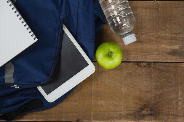 Image depicting school essentials including a backpack, a green apple, a water bottle, a notebook, and a digital tablet on a wooden surface. Ideal for educational content, back-to-school promotions, healthy living articles, and study environment illustrations.