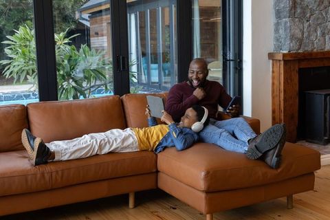 African american father and son relaxing on leather sofa using tablet and phone by pool