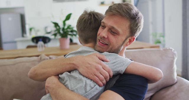 Image shows a joyful father hugging his son on a couch in a modern living room. This image can be used for promoting family values, parent-child relationships, and emotional well-being. It is suitable for advertisements, blogs, and social media posts centered around parenting, family life, and home environments.