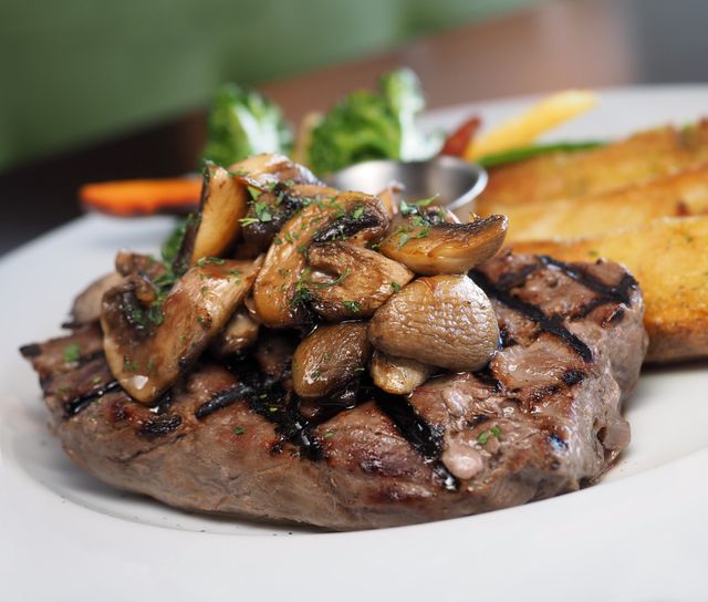 Grilled steak topped with sauteed mushrooms served alongside crispy potato wedges on white plate. Garnished with fresh herbs, presenting gourmet meal perfect for showcasing at a fine dining restaurant, food product advertisement, menu design, or culinary blog.