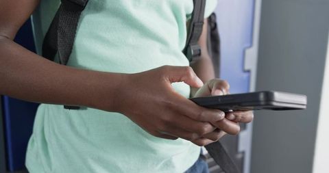 Teen student tapping smartphone in school hallway with backpack straps and lockers