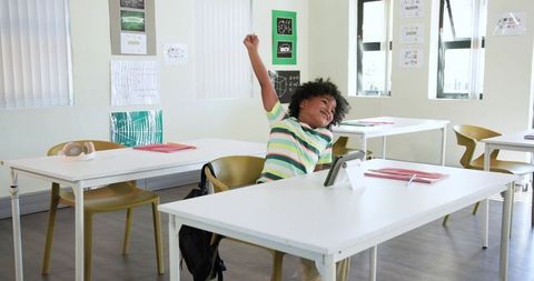 African American Child Raising Hand in Bright Classroom with Tablet and Notebook