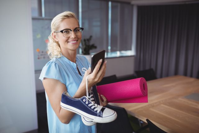 Smiling executive using mobile phone while holding exercise mat and shoes in office