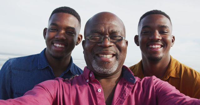 Smiling african american senior father and twin teenage sons standing on a beach taking a selfie. healthy outdoor family leisure time together.