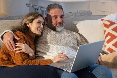 Middle-aged couple cuddling on couch while browsing laptop for cozy evening at home