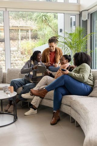Friends laughing on curved sofa sharing tablet and coffee in modern sunlit living room