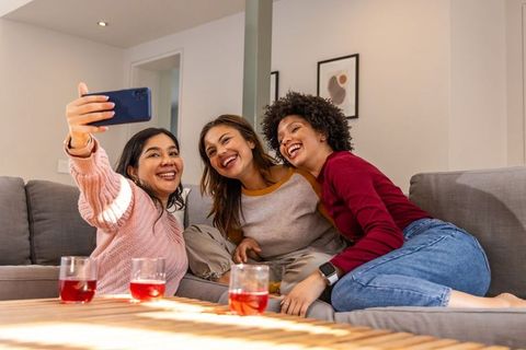 Diverse women taking selfie on sunlit living room sofa, laughing and relaxing