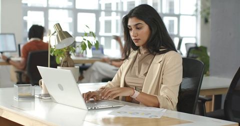 Indian professional typing on laptop in modern open office analyzing charts and focusing