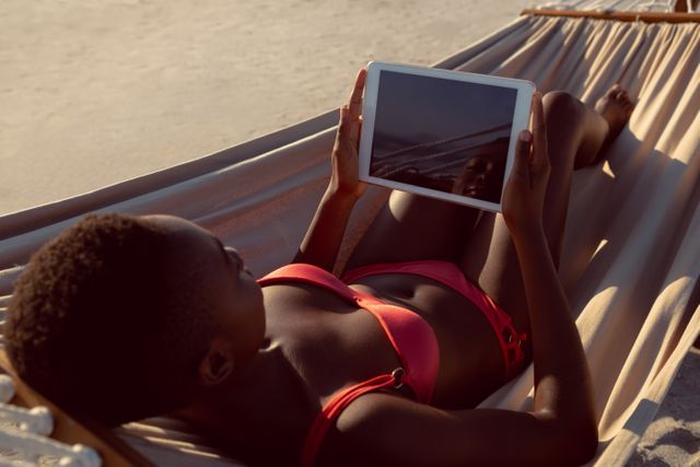 Rear view of young woman African american using digital tablet while relaxing in a hammock on the beach