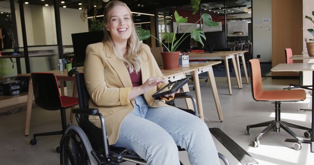 Businesswoman in wheelchair smiling and using digital tablet in a modern office. The inclusive and diverse workplace features contemporary design and casual seating arrangements, highlighting accessibility and utilization of technology. Ideal for showcasing inclusive work environments, technological integration in business, and promoting diversity in the workplace.