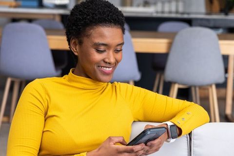 African american woman relaxing on sofa using smartphone and smartwatch in coworking lounge