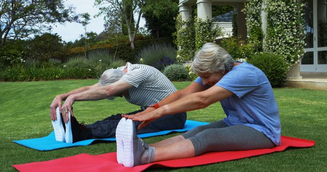Senior couple sitting on yoga mats in garden, stretching toward their toes. Promotes themes of active lifestyle, fitness, and wellness for the elderly. Useful for articles and advertisements related to senior fitness, health routines, and outdoor activities for the elderly.