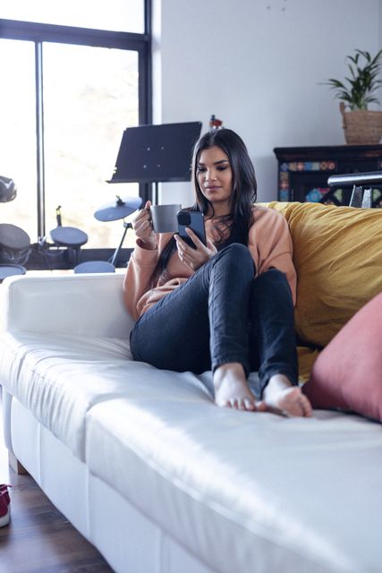 Biracial woman enjoying leisure time at home, sitting comfortably on a sofa with a cup of coffee and using her smartphone. Ideal for concepts related to relaxation, modern lifestyle, home comfort, and technology use in everyday life.