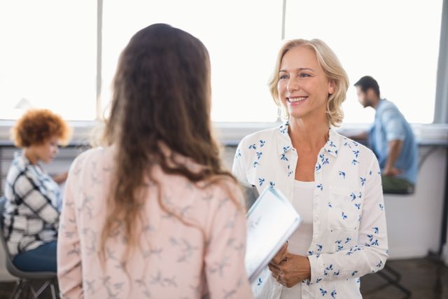 Businesswoman smiling and interacting with female colleague in a modern office environment. Ideal for use in articles or advertisements related to workplace culture, teamwork, professional communication, and business collaboration.