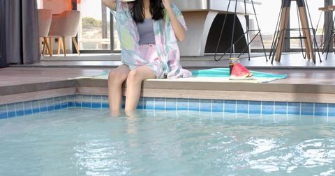Woman sitting at home pool edge wearing pastel cover-up and sun hat with watermelon