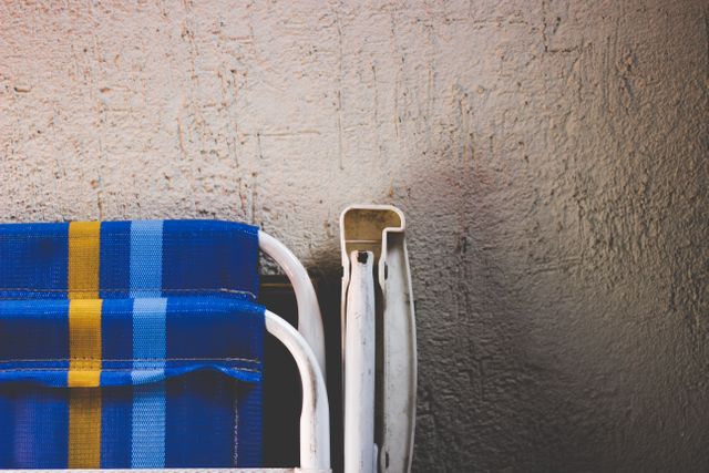 This image depicts a close-up view of a colorful blue and yellow striped outdoor patio chair next to a textured concrete wall. The minimalistic composition emphasizes the simplicity and relaxation of an outdoor setting. The bright, vibrant colors contrast with the neutral, rough surface, which makes it ideal for themes related to outdoor activities, summer relaxation, or modern minimalist designs. Can be used for promotional materials for patio furniture, outdoor products, lifestyle blogs, or seasonal marketing campaigns.