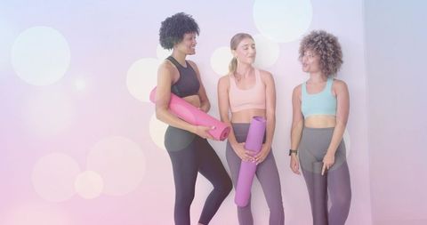 Three women chatting after yoga class holding pink and purple mats in pastel studio