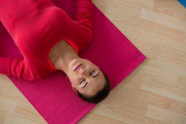 Young woman lying on a pink yoga mat in a yoga studio, appearing relaxed and peaceful. Ideal for use in content related to fitness, wellness, yoga practices, relaxation techniques, and healthy lifestyles. Suitable for blogs, fitness websites, wellness programs, and promotional materials for yoga classes.