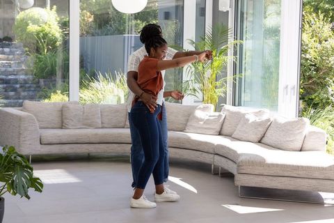 African american couple dancing in sunlit living room with curved sofa and glass walls
