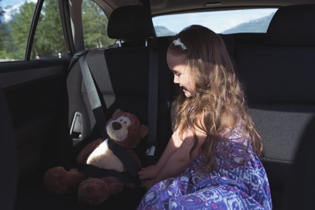 Young girl securing teddy bear with seatbelt in car backseat. Ideal for themes of child safety, family travel, road trips, and childhood innocence. Suitable for use in parenting blogs, travel websites, and safety awareness campaigns.