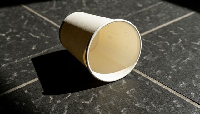 Disposable paper cup lying on dark stone tile casting strong shadow minimal composition