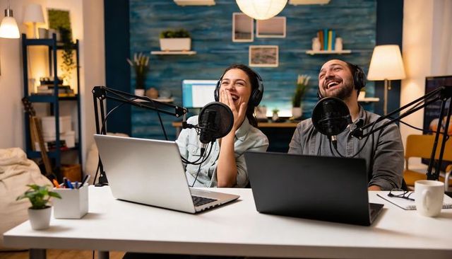 Laughing cohosts recording podcast in cozy home studio with microphones and laptops