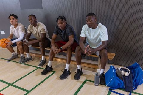 Multiracial male teammates sitting on bench holding basketball and water bottle at gym