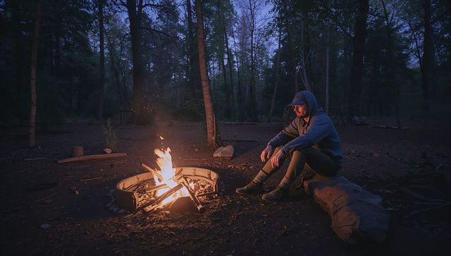 Solo camper sitting by campfire at twilight in quiet forest campsite