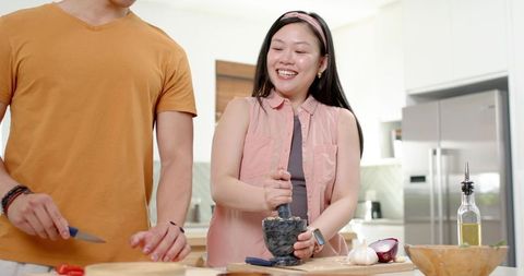 Asian couple preparing meal in modern kitchen, woman grinding garlic with mortar and pestle