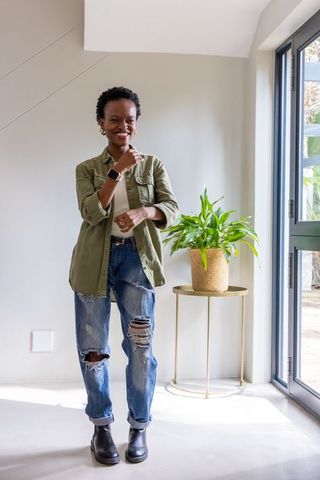 African american woman standing by glass door smiling in bright home wearing olive jacket, ripped je