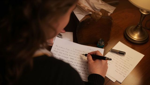 Woman writing with fountain pen at wooden desk with blue inkwell and warm lamp light