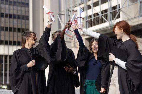 Graduates celebrating success: diverse female students raising diplomas on campus plaza