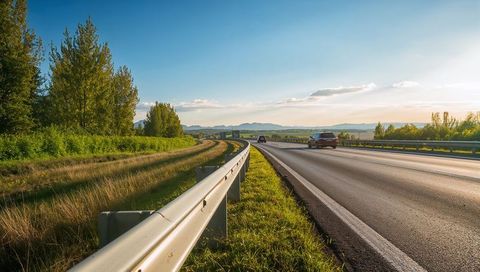 Sunlit guardrail leading along rural highway at golden hour with passing cars, hills