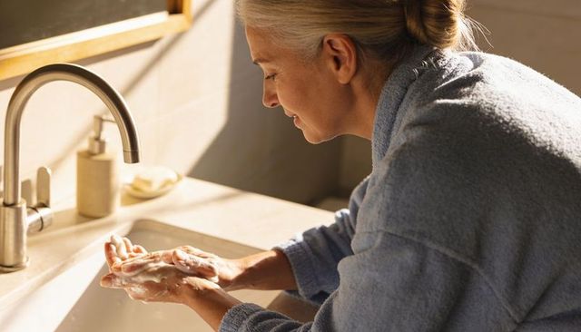 Senior woman washing hands at bathroom sink in blue bathrobe morning hygiene routine