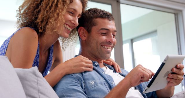 A smiling couple enjoying time together at home while using a digital tablet on a comfortable couch. This image is ideal for promoting home technology products, online services, and advertisements depicting modern lifestyles. It also works well for campaigns focused on family bonding, relaxation, and digital connectivity.