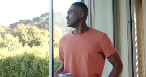 African American man holding mug at sliding door, enjoying morning garden view
