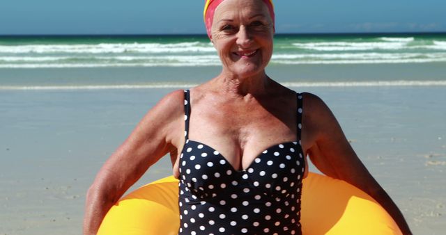 Senior woman in polka dot swimsuit smiling and holding inflatable ring at beach. Perfect for content related to senior lifestyle, beach activities, retirement, and health and wellness during summer. Highlights aging with joy and energy.
