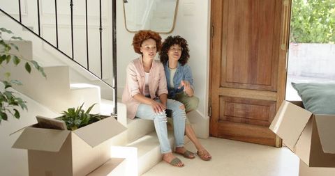 Two diverse women sitting on staircase unpacking moving boxes in bright modern entryway