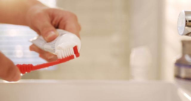 Close-up view focusing on hands squeezing toothpaste onto a red toothbrush. Ideal for promoting dental hygiene, morning routine tips, or personal care products. This can be used in advertisements or educational materials about the importance of dental care and maintaining healthy habits.