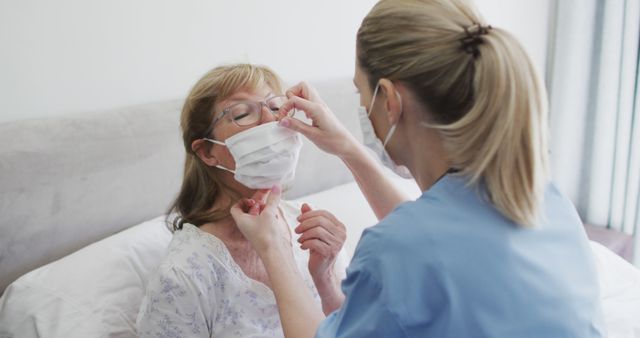 Caregiver in blue uniform adjusting a white facemask for elderly woman sitting on bed. Useful for topics related to elderly care, healthcare services, emotional support, nursing, and medical assistance at home or in hospital. Could be used in articles, blogs, or advertisements focused on senior health and caregiving.