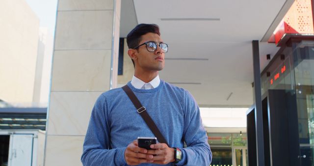 Young professional man standing outdoors, using smartphone while wearing glasses and messenger bag. Suitable for content about urban lifestyle, modern technology use, professional life, and young adults.