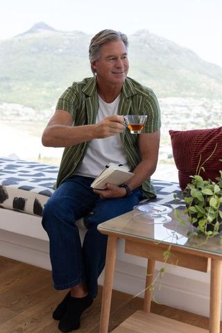 Mature man sipping tea while journaling on sunlit window bench with mountain view