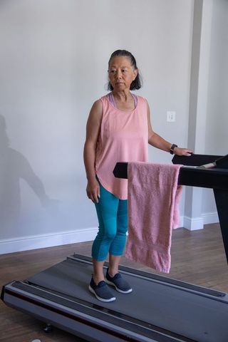 Senior asian woman standing on treadmill checking smartwatch while holding pink towel