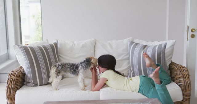 A girl laying on a comfortable sofa plays affectionately with her small dog. The bright and cozy living room setting evokes feelings of home, leisure, and the special bond between children and pets. Great for themes of family life, pet care, children at home, and interior design.