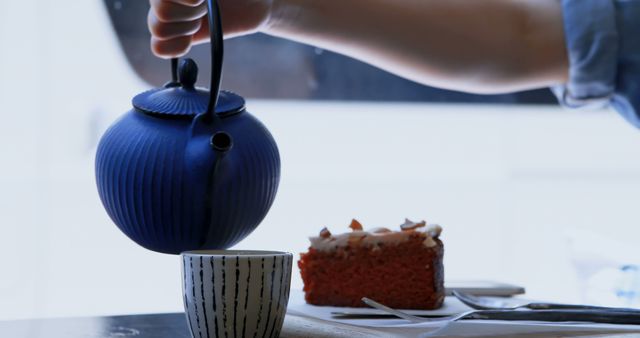 A person is pouring tea from a blue teapot into a cup, with a slice of red velvet cake on the side, with copy space. Capturing a moment of relaxation, the image evokes a sense of comfort and indulgence during a tea break.