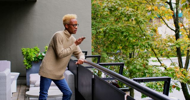 A man with glasses and a sweater is standing on a balcony, speaking on a smartphone. The balcony overlooks trees with autumn foliage. Use this for promotions about modern living, outdoor communication, relaxation, or technology. Suitable for blogs, advertisements, or lifestyle magazines discussing city life and personal connectivity.