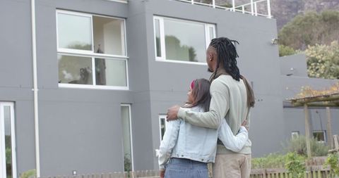 Interracial couple embracing in front of modern gray suburban house celebrating new homeownership